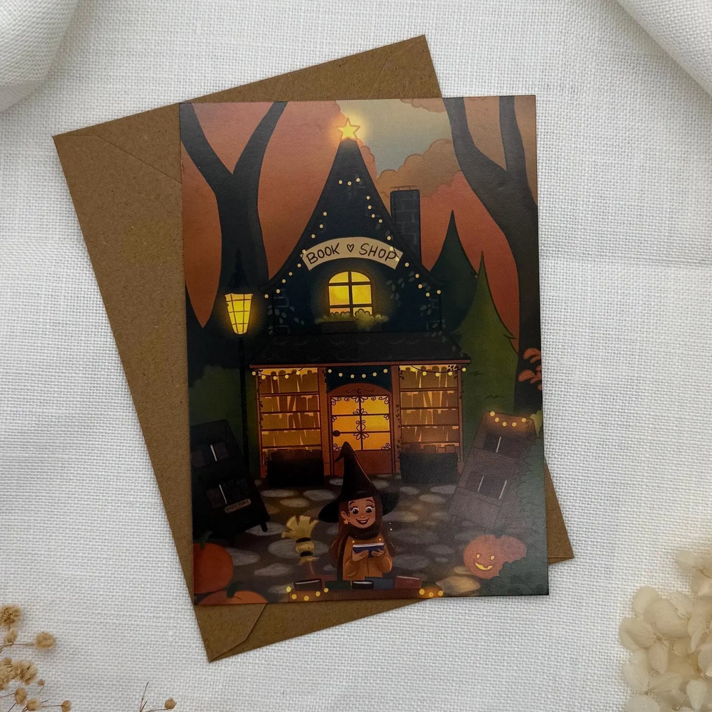 Postkarte „Bookshop Witch“ Herbstliche Hexe vor Buchladen | Magische Herbstkarte mit Kürbis & Lichterglanz